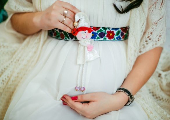 Une femme enceinte tient une petite poupée près de son ventre, portant une robe blanche et une ceinture brodée, avec des Bijoux de Grossesse subtils aux doigts.