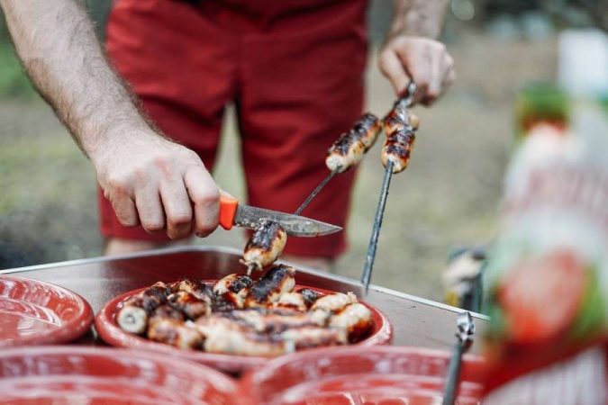 Un homme en short rouge joue les chefs au camping en retirant des saucisses grillées d'une brochette avec un couteau au-dessus d'une assiette.