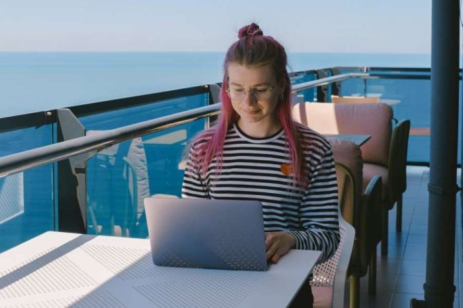 Une jeune femme travaille sur son ordinateur portable, incarnant le quotidien d'un nomade digital sur un balcon avec vue sur la mer.