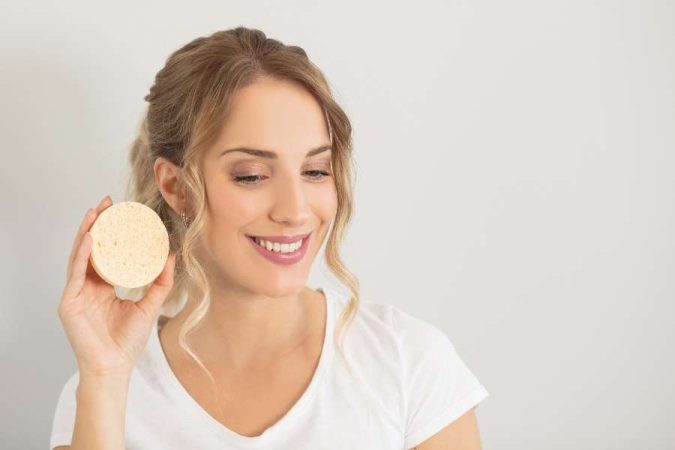 Une femme blonde sourit en montrant une éponge douce pour éliminer les impuretés de la peau.