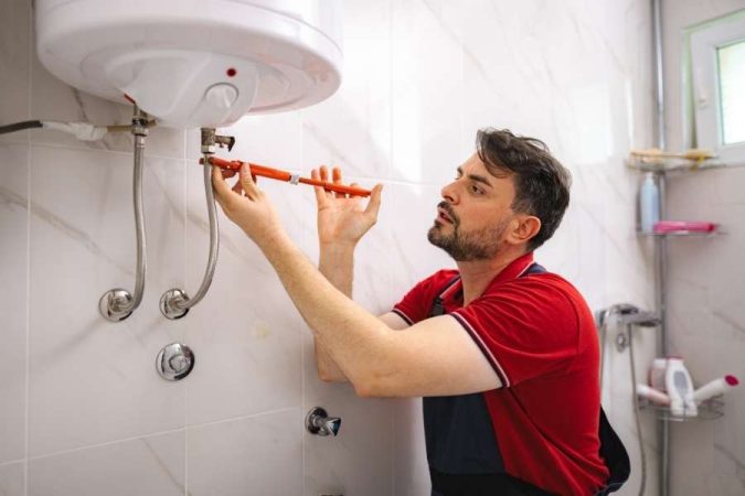 Un plombier en tenue de travail rouge utilisant une clé à molette pour réparer un petit chauffe-eau électrique fixé au mur blanc d'une salle de bains.